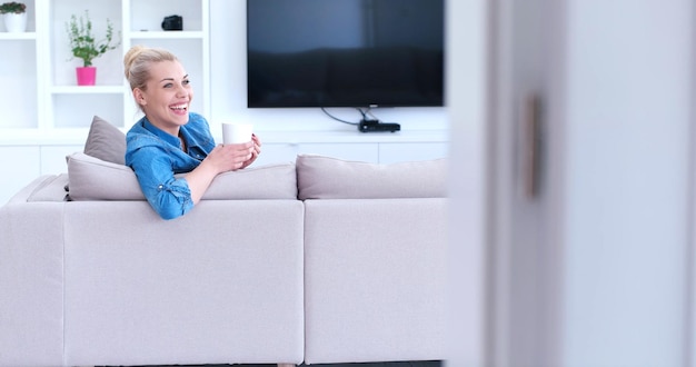 Schöne blonde Frau, die eine Tasse Kaffee genießt, während sie auf einem Sofa in ihrer Wohnung sitzt