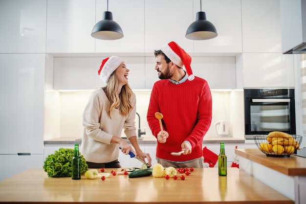 Schöne blonde Frau, die Gurke schneidet, während stehend und Mann vorgibt, Trommeln mit Mischlöffeln zu spielen. Beide haben Weihnachtsmützen auf den Köpfen