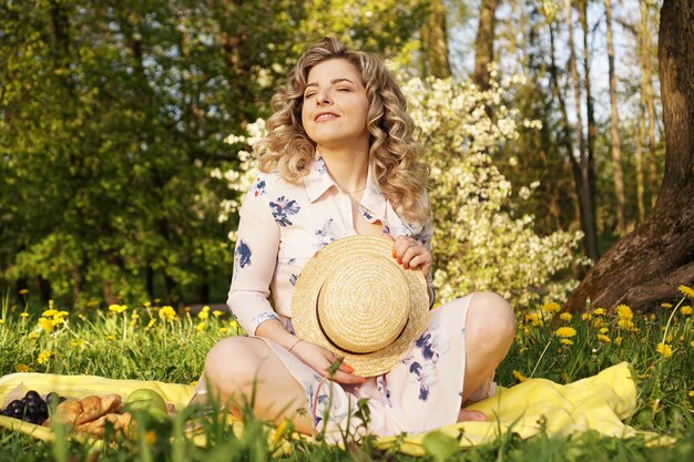 Schöne blonde Frau - glücklicher Lebensstil, Wochenende für einen Spaziergang in einem Picknickpark im Sommergarten, Modell sitzt auf Plaid mit Essen - Sommerwetter