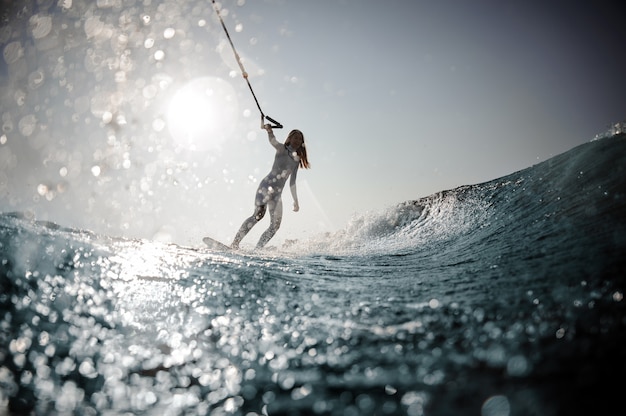 Schöne blonde Frau im weißen Badeanzug, die auf dem Wakeboard steht und ein Seil auf den beugenden Knien hält