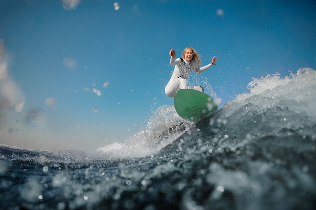 Schöne blonde Frau im weißen Sportbadeanzug reitet auf dem grünen Wakeboard auf den beugenden Knien