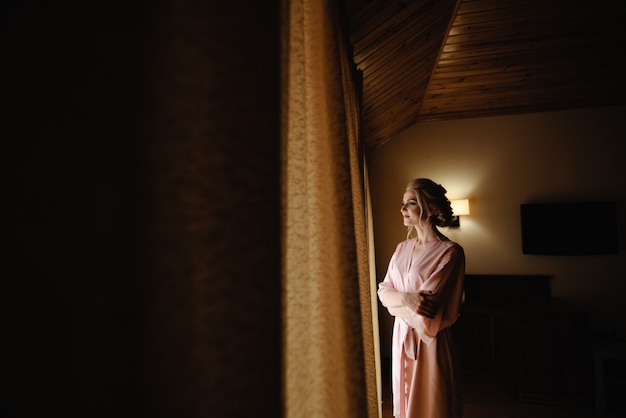 Schöne blonde Frau in einem rosa Bademantel, der auf das Fenster schaut