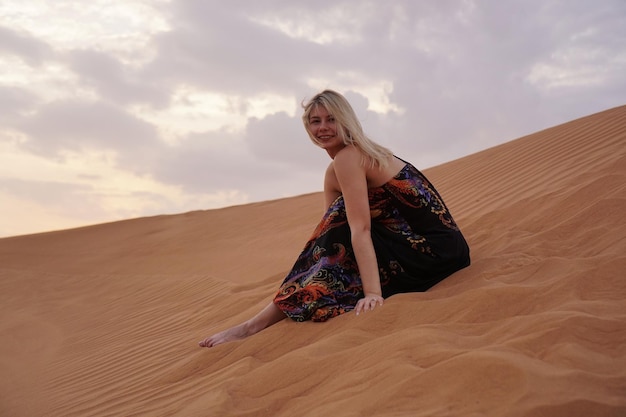 Schöne blonde Frau in einem schwarzen Kleid sitzt auf dem heißen Sand in der Wüste