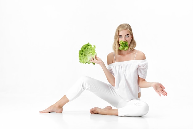 Schöne blonde Frau in einer weißen Bluse, die einen frischen grünen Salat auf einem weißen Hintergrund isst. Gesundheit und Ernährung