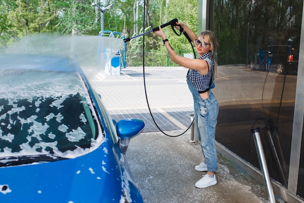 Schöne blonde Frau in Jeans-Overalls wäscht das Auto