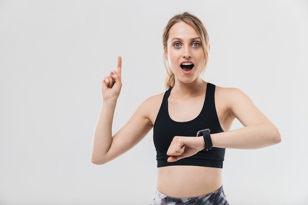 Schöne blonde Frau in Sportkleidung, die während der Fitness im Fitnessstudio isoliert über weißer Wand trainiert und auf die Armbanduhr schaut