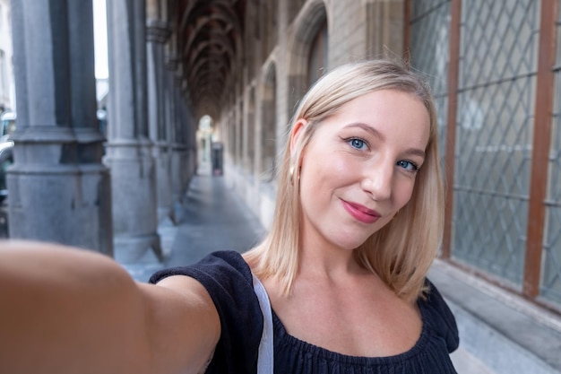 Schöne blonde Frau in stilvollen Kleidern macht ein Selfie, während sie vorbei an einem alten Gebäude läuft