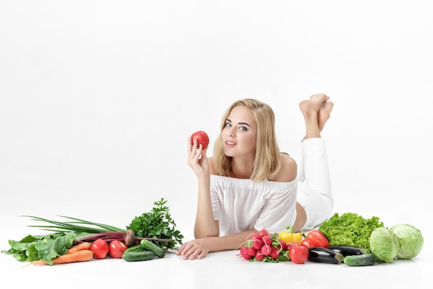 Schöne blonde Frau in weißen Kleidern und viel frischem Gemüse auf einem weißen Hintergrund. Mädchen, das eine Tomate hält