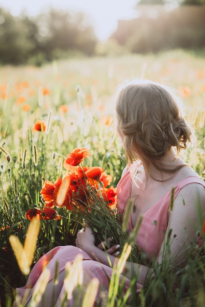 Schöne blonde Frau mit einem Blumenstrauß von Mohnblumen sitzt in einem Feld im Sommer an einem sonnigen Tag.