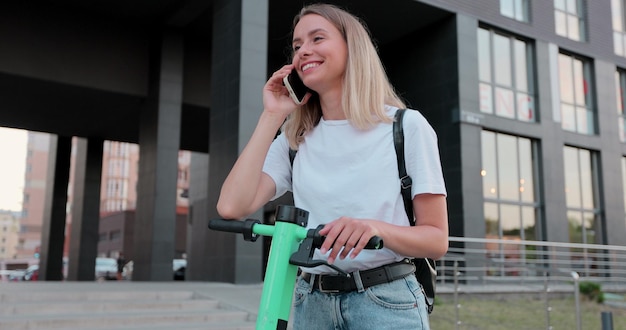 Schöne blonde Frau mit einem Elektroroller auf der Straße macht Halt, um mit jemandem auf dem Smartphone zu chatten