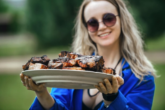 schöne blonde frau mit einem großen teller gegrilltem fleisch zum grillen in der natur bei einem picknick