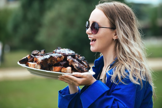 Schöne blonde Frau mit einem großen Teller mit fertig gebratenem Fleisch zum Grillen in der Natur