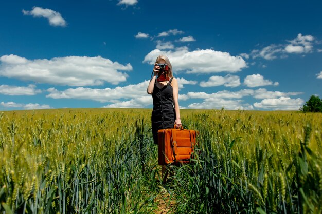 Schöne blonde Frau mit Koffer und Weinlesekamera im Weizenfeld am sonnigen Tag