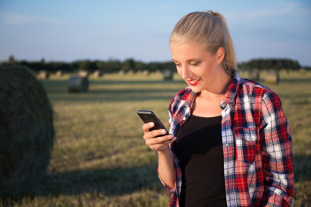 Schöne blonde Frau mit Mobiltelefon im Feld mit Heuhaufen