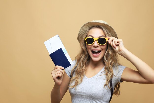 Schöne blonde Frau mit Sonnenbrille posiert mit Pass mit Tickets über beigefarbenem Hintergrund.