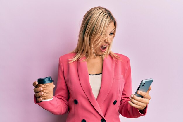 Schöne blonde Frau mittleren Alters, die Smartphone benutzt und eine Tasse Kaffee trinkt, wütend und wütend, schreit frustriert und wütend und schreit vor Wut. Wut und aggressives Konzept.