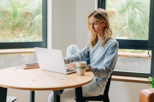 Schöne blonde Frau sitzt am Tisch und arbeitet online an einem Laptop