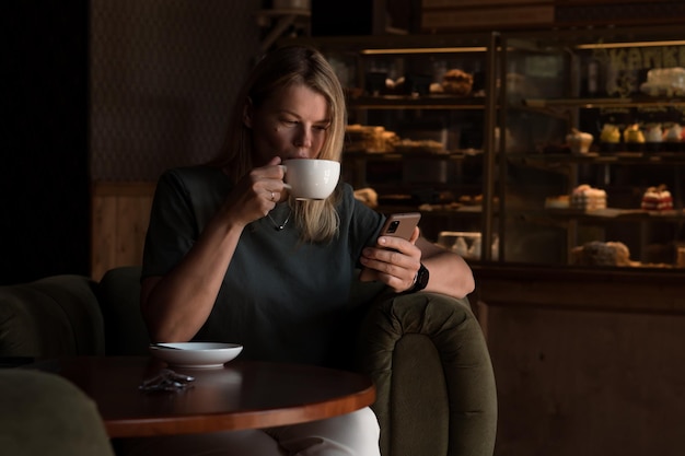 Schöne blonde Frau sitzt im Kaffeehaus und in der Bäckerei und trinkt Tee oder Kaffee und benutzt ein Smartphone