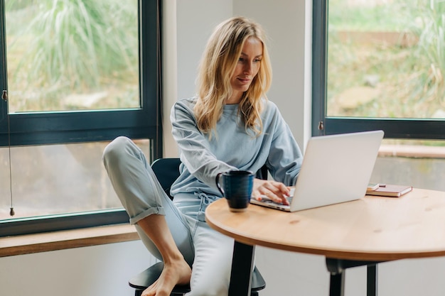 Schöne blonde Frau sitzt zu Hause im Wohnzimmer am Tisch und arbeitet aus der Ferne an einem Laptop