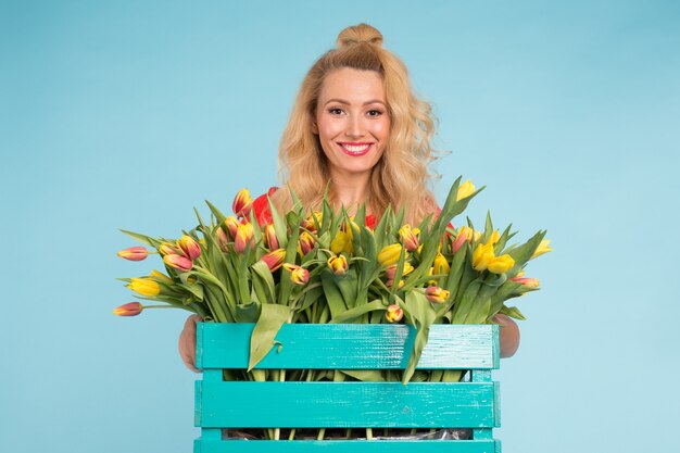 Schöne blonde Gärtnerin, die Box mit Tulpen auf Blau hält