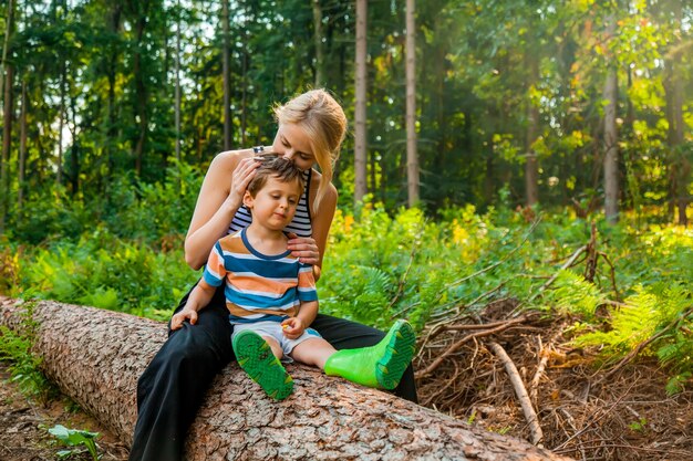 Schöne blonde Haarmutter sitzt mit Sohn im Wald auf Baumstamm