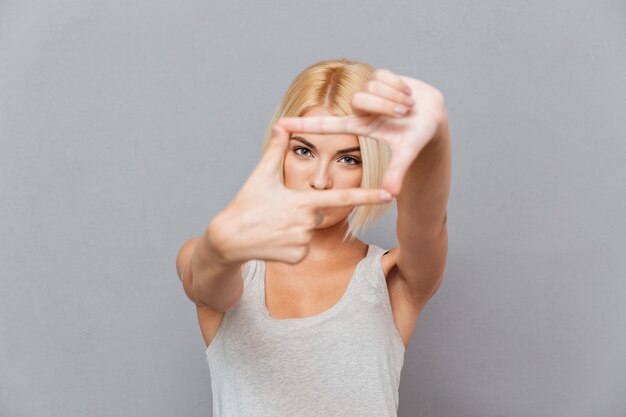 Schöne blonde junge Frau, die Rahmen mit den Fingern über grauer Wand macht