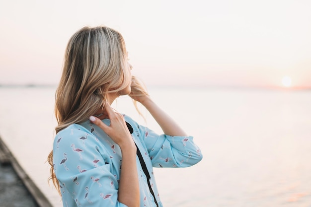 Schöne blonde junge Frau im blauen Kleid auf dem Pier und Blick auf den Sonnenuntergang