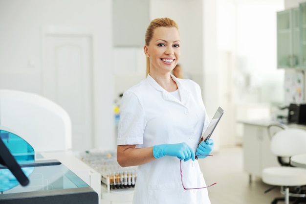 Schöne blonde Laborassistentin im weißen Kittel und in den Gummihandschuhen, die im Labor mit Tablette in ihren Armen stehen.