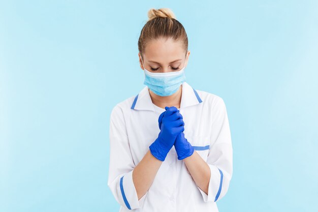 Schöne blonde Ärztin in Uniform und Maske, die isoliert über blauer Wand steht und für viel Glück betet