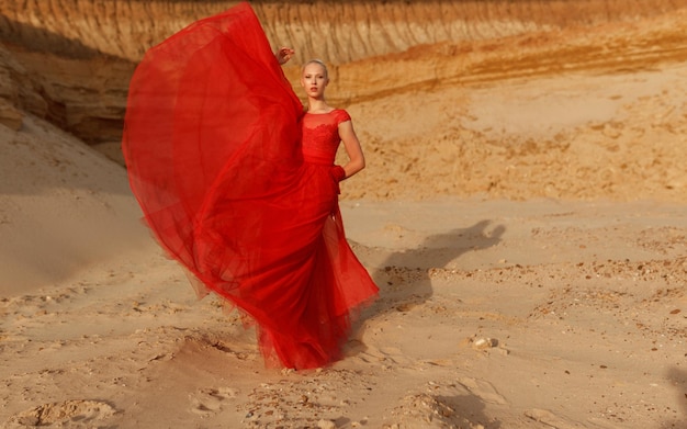 Schöne blonde weibliche Modellabnutzung im roten Kleid in der Wüste, Kamera betrachtend. Porträt in voller Länge.