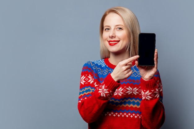 Schöne Blondine im Weihnachtspullover mit Handy auf grau