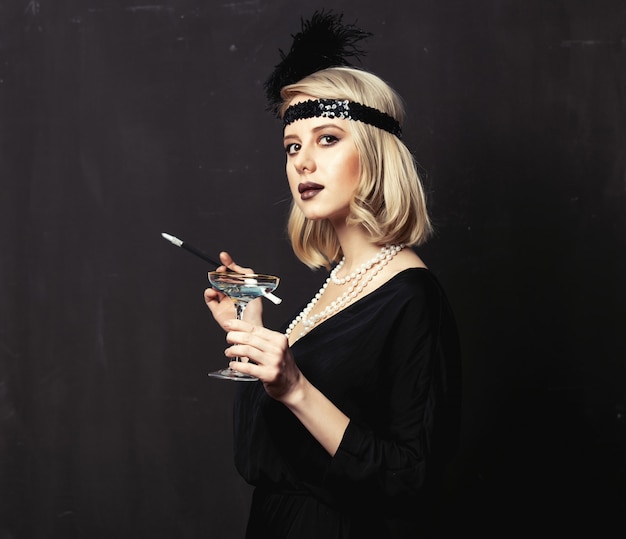 Schöne Blondine in den Zwanziger Jahren kleiden mit Pfeife und Cocktail auf dunklem Hintergrund