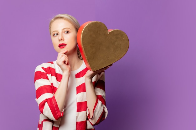 Schöne Blondine in der roten Jacke mit Herzform-Geschenkbox auf lila Hintergrund