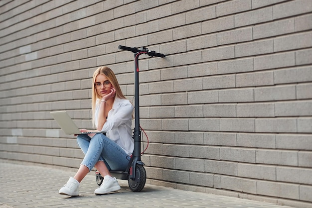 Schöne Blondine in Freizeitkleidung, die tagsüber auf Elektrorollern im Freien sitzt