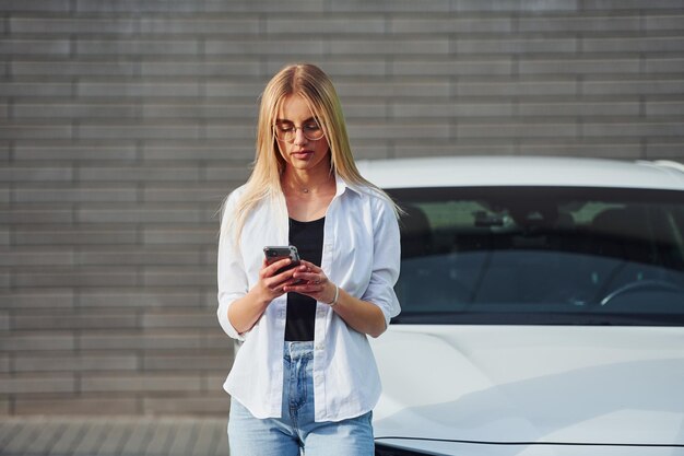 Schöne Blondine in Freizeitkleidung mit Telefon in der Nähe eines weißen Autos ist tagsüber im Freien