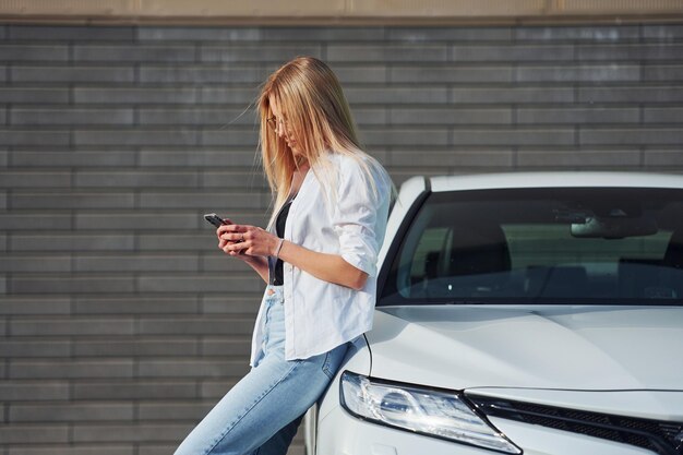 Schöne Blondine in Freizeitkleidung mit Telefon in der Nähe eines weißen Autos ist tagsüber im Freien