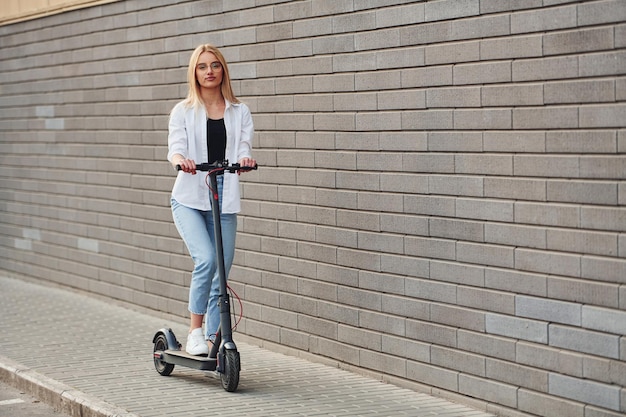 Schöne Blondine in legerer Kleidung, die tagsüber bei sonnigem Wetter im Freien mit Elektroroller fährt