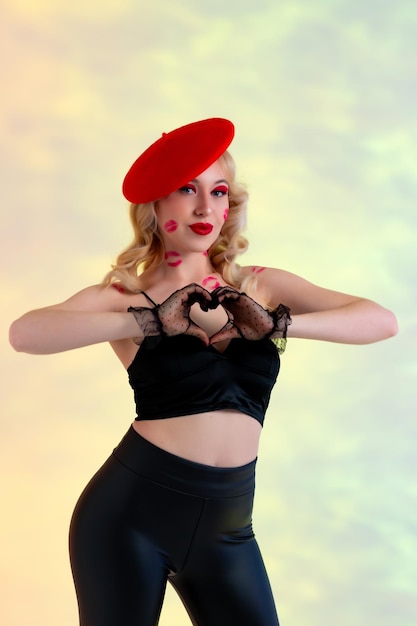 Schöne Blondine in schwarzen Lederhosen und rotem Hut, schwarzen Spitzenhandschuhen. Spuren von Küssen auf Körper und Gesicht. Zeigt Herz mit Händen. Gelber abstrakter Hintergrund