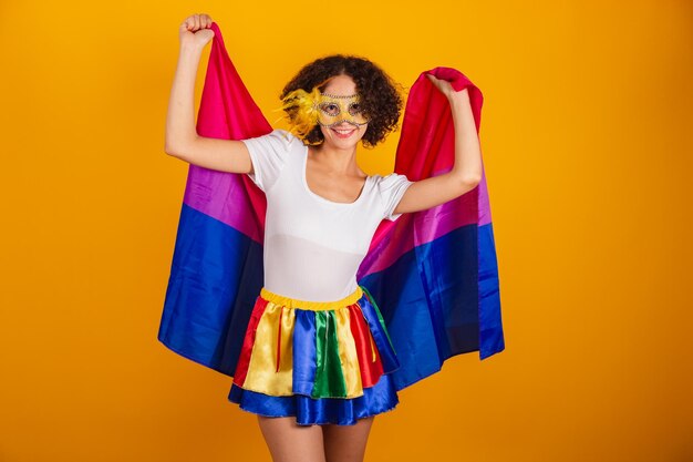 Schöne Brasilianerin in Karnevalskleidung, buntem Rock und weißem Hemd, die eine Maske mit bisexueller Flagge trägt