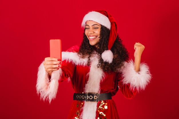 Schöne brasilianische schwarze Frau, die als Weihnachtsmann-Weihnachtskleidung gekleidet ist und mit etwas auf dem Smartphone feiert