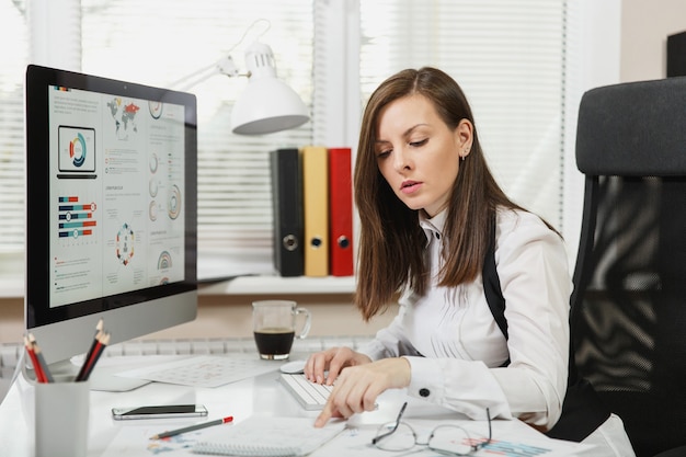 Schöne braunhaarige Geschäftsfrau in Anzug und Brille, die mit einer Tasse Kaffee am Schreibtisch sitzt, am Computer mit Dokumenten im hellen Büro arbeitet und in die Kamera schaut