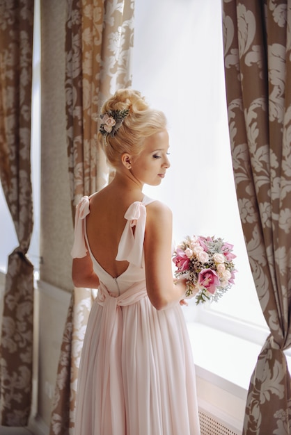 Schöne Braut der jungen Frau mit Hochzeitsblumenstrauß von den Rosen und von Orchideen, die nahes Fenster stehen