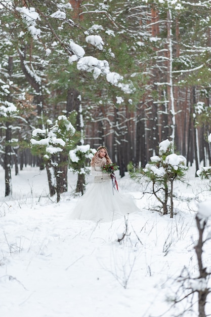 Schöne Braut, die mit Blumenstrauß im verschneiten Wald aufwirft. Winterhochzeit. Kunstwerk. Porträt in voller Länge