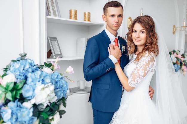 Schöne Braut im weißen Kleid und im Bräutigam im Anzug, posierend im weißen Studio-Interieur, Hochzeit