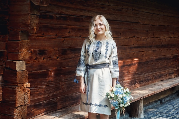 Schöne Braut in einem bestickten Hemd mit einem Blumenstrauß auf dem Hintergrund eines Holzhauses.