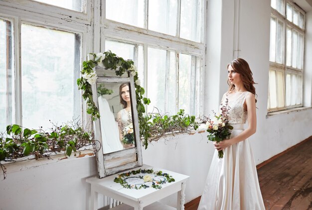 Schöne Braut in einem Hochzeitskleid in einem hellen, stilvollen Studio Hochzeit im europäischen Stil Dekor und Blumenstrauß in der Hand