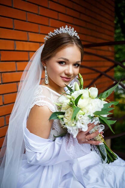 Schöne Braut in Schleier und weißem Mantel sitzt auf einer Decke mit einem Hochzeitsstrauß in den Händen