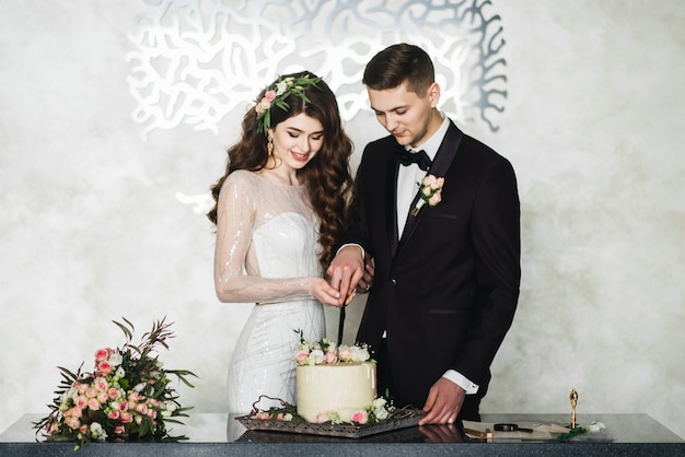 Schöne Braut und Bräutigam, die einen Wunsch machen, während sie stehen, die Hochzeitstorte zusammen schneiden