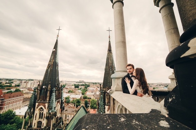 Schöne Braut und stilvoller Bräutigam umarmen sich auf dem Balkon der alten gotischen Kathedrale mit Panoramablick auf die Stadt