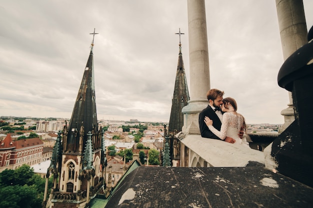 Schöne Braut und stilvoller Bräutigam umarmen sich auf dem Balkon der alten gotischen Kathedrale mit Panoramablick auf die Stadt